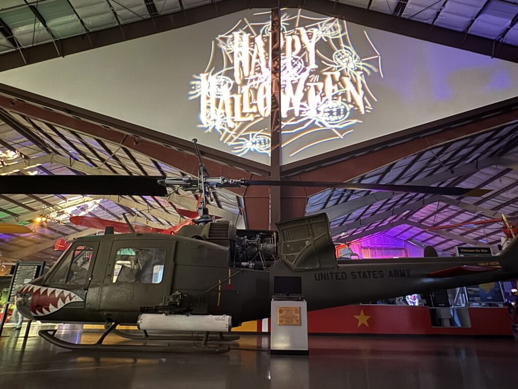 A Vietnam-era Army helicopter with a shark-mouth nose on display in the museum hangar, with a Happy Halloween spider web projection overhead