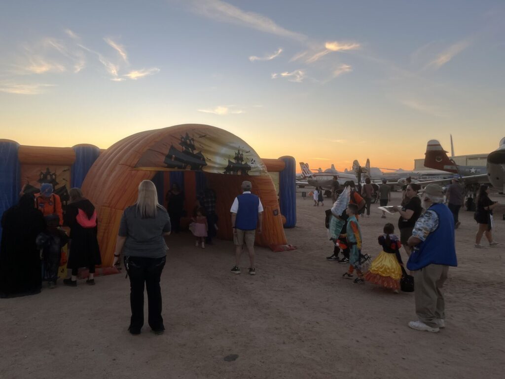 Children in costumes and volunteers at an inflatable trick-or-treat tunnel at sunset, with aircraft in the background